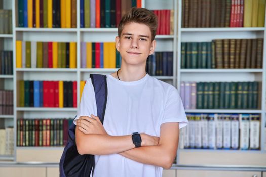 Single portrait of smiling confident male student teenager with backpack with crossed arms looking at camera in the library. School, education, knowledge, adolescence concept