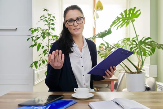 Business woman looking at camera talking, holding papers documents in hands, home interior background. Online consultation, remote work, paperwork, internet, technologies in business, education