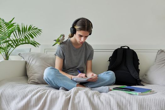 Male teenager with pet green parrot quaker on his shoulder, guy student sitting in headphones with textbooks notebooks books at home on bed