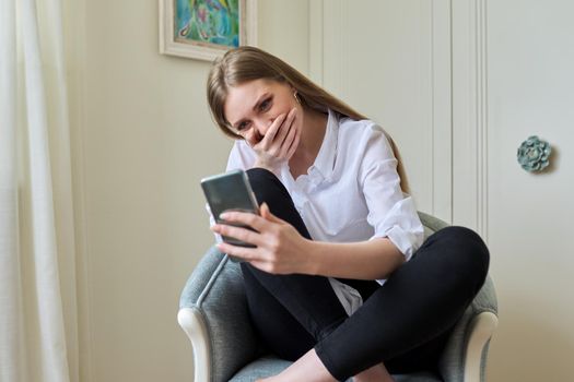 Emotional laughing young beautiful woman looking at smartphone screen, relaxing sitting at home in chair. Video call, emotional communication, joy, happiness, surprise, technology in everyday life