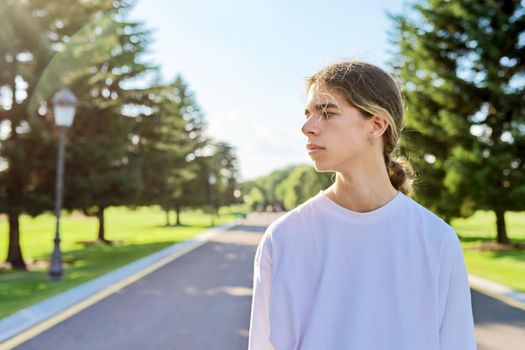 Portrait of handsome teenage guy, profile view outdoors, copy space. Serious hipster teenager with long hair wearing white t-shirt on road on sunny summer day. Youth, adolescence, people, lifestyle concept