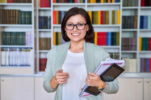 Portrait of positive smiling middle aged woman teacher, psychologist, mentor, therapist, counselor, business woman looking at camera. Female in glasses with textbooks tablet, library office with books