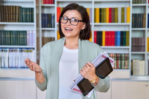 Portrait of positive smiling middle aged woman teacher, psychologist, mentor, therapist, counselor, business woman looking at camera. Female in glasses with textbooks tablet, library office with books