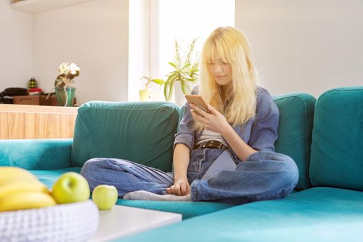 Smiling happy female teenager 16 years old sitting at home on couch looking at smartphone screen, texting, browsing blog, social networks, chatting with friends online