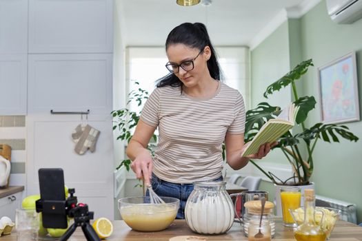 Woman preparing apple pie at home in kitchen with recipe book in her hand, looking in smartphone webcam, talking online using video call. Female food blogger recording video recipe