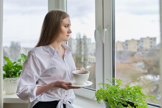 Portrait of young beautiful woman in white shirt with cup of coffee near window. Blonde female relaxing looking out the window of winter autumn city