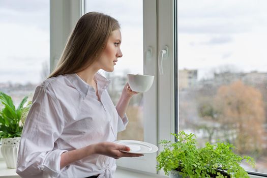 Portrait of young beautiful woman in white shirt with cup of coffee near window. Blonde female relaxing looking out the window of winter autumn city