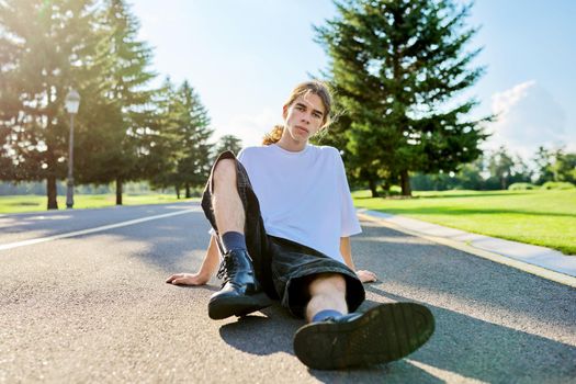 Fashion portrait of hipster teenager guy sitting on road. Serious posing young male with long hair looking in camera,in white t-shirt shorts boots on summer day. Youth, adolescence, people, lifestyle