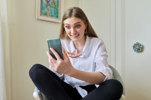 Emotional laughing young beautiful woman looking at smartphone screen, relaxing sitting at home in chair. Video call, emotional communication, joy, happiness, surprise, technology in everyday life