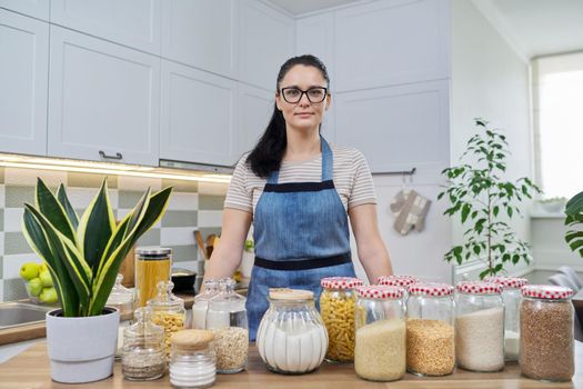 Portrait of smiling woman housewife in an apron in kitchen. Jars of cereals on the table, organizing and storing food