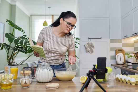 Woman preparing apple pie at home in kitchen with recipe book in her hand, looking in smartphone webcam, talking online using video call. Female food blogger recording video recipe