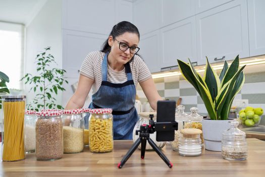 Online broadcast, blog about food, woman in an apron with jars of cereals noodles flour, looking at smartphone camera, telling and showing about food storage, organizing kitchen