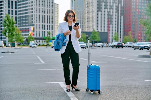 Middle aged businesswoman with suitcase talking on mobile phone in outdoor parking, urban style, city architecture background. Business travel trip, business people of mature age, success, modern city