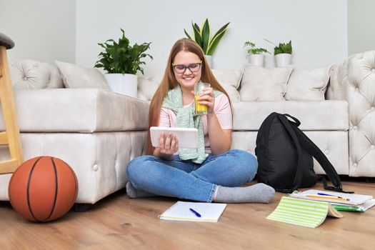 Teenage girl sitting at home on floor looking at screen of digital tablet, having rest, drinking orange juice. Teenager lifestyle, home, vacation