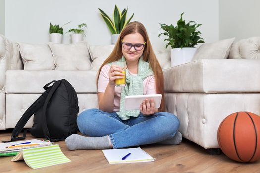Teenage girl sitting at home on floor looking at screen of digital tablet, having rest, drinking orange juice. Teenager lifestyle, home, vacation