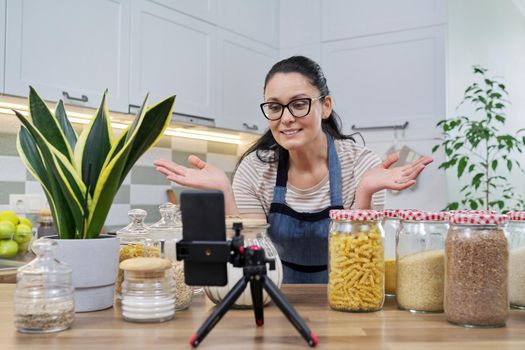 Online broadcast, blog about food, woman in an apron with jars of cereals noodles flour, looking at smartphone camera, telling and showing about food storage, organizing kitchen