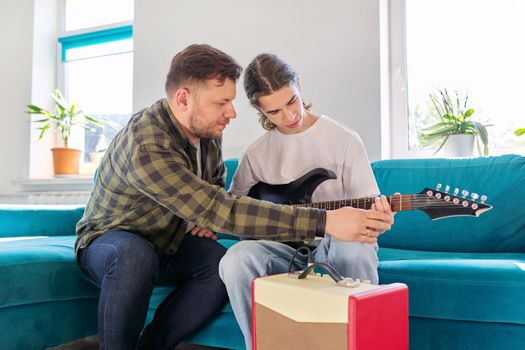 Father teaches his teenage son to play the electric guitar. Family sitting together at home in living room on couch. Parent-child relationship, lifestyle, creativity, music, teenagers concept