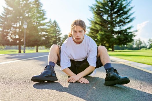 Fashion portrait of hipster teenager guy sitting on road. Serious posing young male with long hair looking in camera,in white t-shirt shorts boots on summer day. Youth, adolescence, people, lifestyle