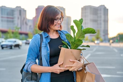 Middle aged woman with paper shopping bags with buying plant, outdoor, store parking lot background. Smiling female shopping in city using craft recycled brown bags, ficus lirata in hand, green trends