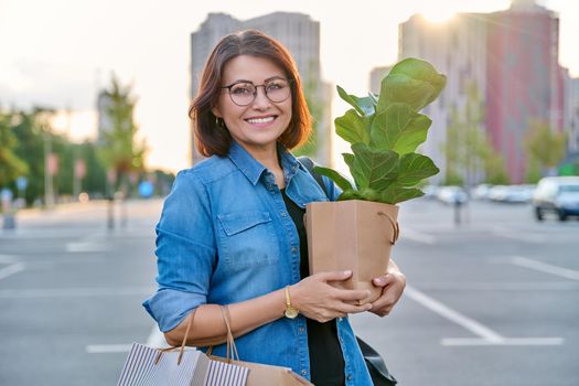 Middle aged woman with paper shopping bags with buying plant, outdoor, store parking lot background. Smiling female shopping in city using craft recycled brown bags, ficus lirata in hand, green trends