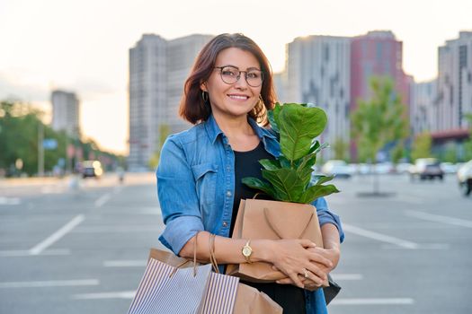 Middle aged woman with paper shopping bags with buying plant, outdoor, store parking lot background. Smiling female shopping in city using craft recycled brown bags, ficus lirata in hand, green trends