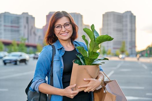 Middle aged woman with paper shopping bags with buying plant, outdoor, store parking lot background. Smiling female shopping in city using craft recycled brown bags, ficus lirata in hand, green trends