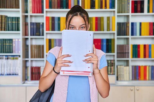 Single portrait of smiling female teenage schoolgirl looking at camera with blank notebook and place for text, library interior, books background