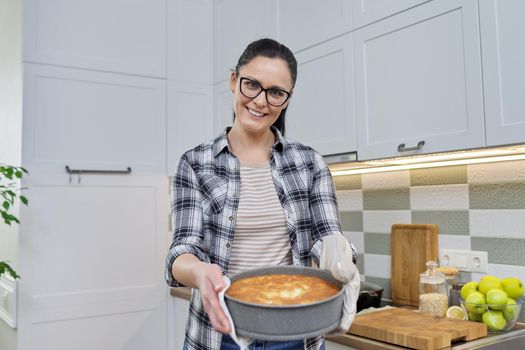 Smiling woman in kitchen mittens with hot freshly baked pie, kitchen interior background