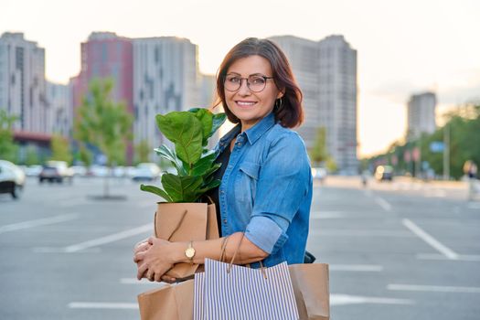 Middle aged woman with paper shopping bags with buying plant, outdoor, store parking lot background. Smiling female shopping in city using craft recycled brown bags, ficus lirata in hand, green trends