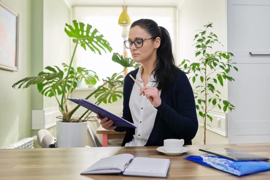 Business woman looking at camera talking, holding papers documents in hands, home interior background. Online consultation, remote work, paperwork, internet, technologies in business, education