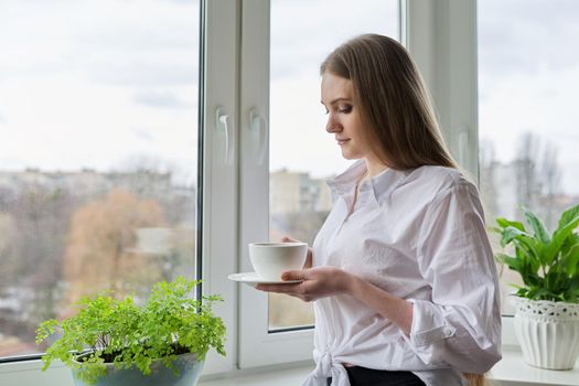 Portrait of young beautiful woman in white shirt with cup of coffee near window. Blonde female relaxing looking out the window of winter autumn city