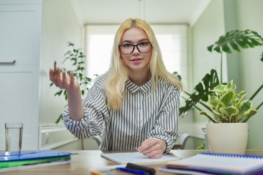 Portrait of teenager female student looking at camera, talking smiling, sitting at home at the table with notebooks textbooks. Looks in webcam, online education, video lesson, conference, e-learning