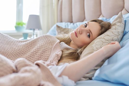 Young beautiful smiling blonde woman resting in bed at home. Close up face looking at camera, blue bed under pink pastel knitted blanket