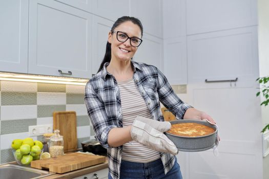 Smiling woman in kitchen mittens with hot freshly baked pie, kitchen interior background