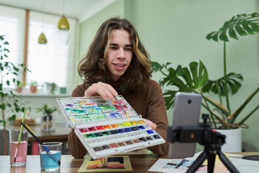 Online learning, drawing, e-education. Teenage guy drawing with watercolors, watching video class on smartphone, sitting at table at home. Creativity, technology, hobby, teens concept