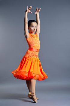 Child girl in an orange sports dress posing in dance movement on gray studio background, sports dances ballroom and latin american for children and adolescents