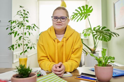 Portrait of girl 12, 13 years old in yellow sweatshirt with glasses looking at the camera sitting at home at table