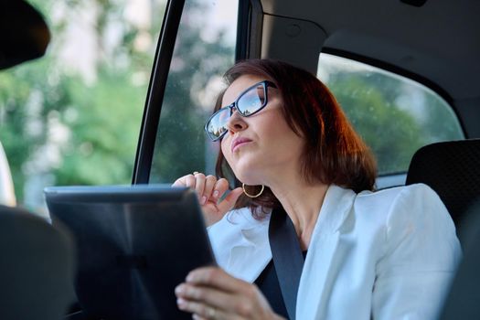 Middle-aged businesswoman in car in passenger backseat. Serious female in glasses with digital tablet, tired woman looking out window. Business people of mature age, city, fatigue frustration