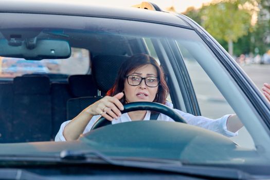 Middle-aged woman driver with glasses driving car, female is emotional, outraged, angry, upset. Mature people, transportation, lifestyle concept