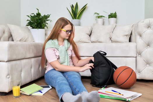 Lifestyle, education children 12, 13 years old concept. Teenage girl sitting at home on the living room floor with backpack, books, school notebooks, basketball