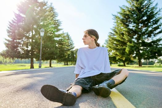 Portrait of handsome teenage guy, profile view outdoors, copy space. Serious hipster teenager with long hair wearing white t-shirt on road on sunny summer day. Youth, adolescence, people, lifestyle concept