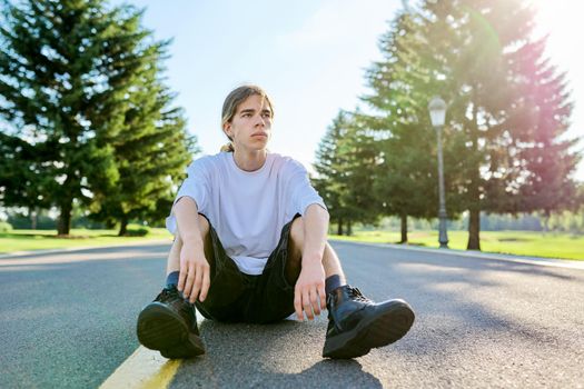 Fashion portrait of hipster teenager guy sitting on road. Serious posing young male with long hair wearing white t-shirt shorts boots on sunny summer day. Youth, adolescence, people, lifestyle concept