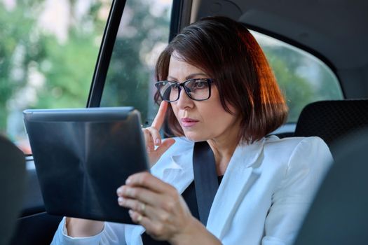 Middle-aged business woman in car in passenger backseat. Serious female in glasses with digital tablet. Business people of mature age, city, emotion fatigue meditation sadness depression frustration