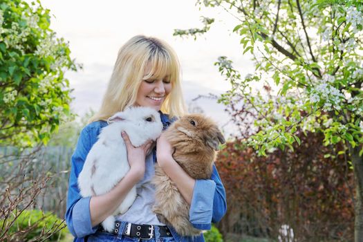 Beautiful smiling girl teenager with a couple of decorative rabbits in their hands. Blonde female in denim clothes with two bunnies in a spring blooming garden