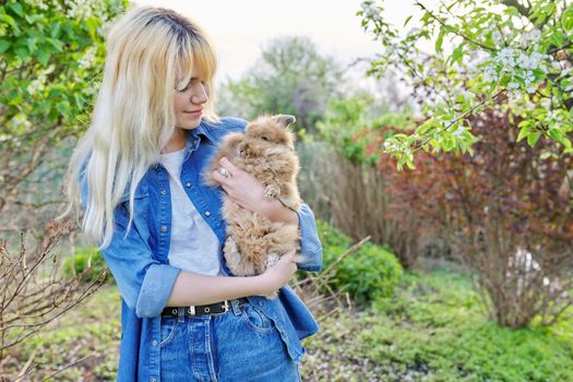 Young blonde woman with decorative rabbit in the spring garden. Female teenager with a pet fluffy bunny. Nature, pets, domestic animals, copy space
