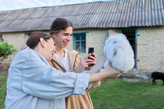 Mom and her teenage son laugh, photograph a decorative rabbit fluffy white bunny on a smartphone, garden farm background. Country life, family, mother and teen son
