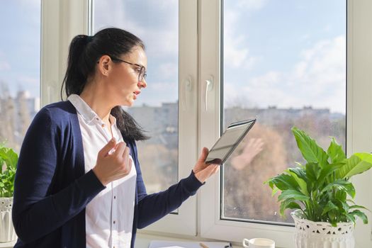 Serious business woman working remotely in her home office, talking on video call, using digital tablet. Freelance, technology, telecommunications, business remotely, video communication near window