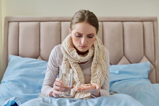 Sick woman with glass of water and pills in her hand, young female sitting at home in bed taking medicine. Medicine, pharmacology, flu season, seasonal diseases, people concept