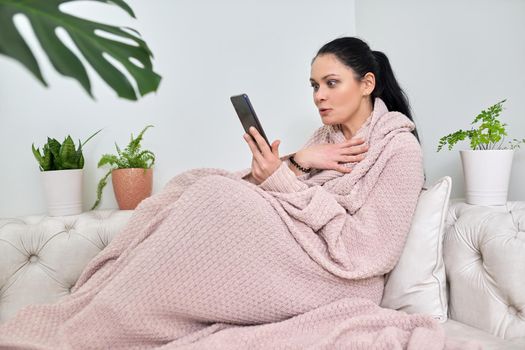 Surprised emotional middle-aged woman using smartphone for video call, female in autumn winter season under warm knitted blanket sitting on sofa in living room. Video, online, virtual meetings, communication