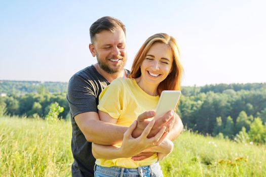 Beautiful happy adult couple taking selfie on smartphone, sunny summer day meadow forest sky background. Relationships, happiness, relaxation, love, people 30s 40s age